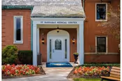 Entrance to St Barnabas Medical Center with a white door under a small covered porch. The building is made of red brick with windows on either side. There are flower beds with red and white flowers and a bench on the right side near the walkway.