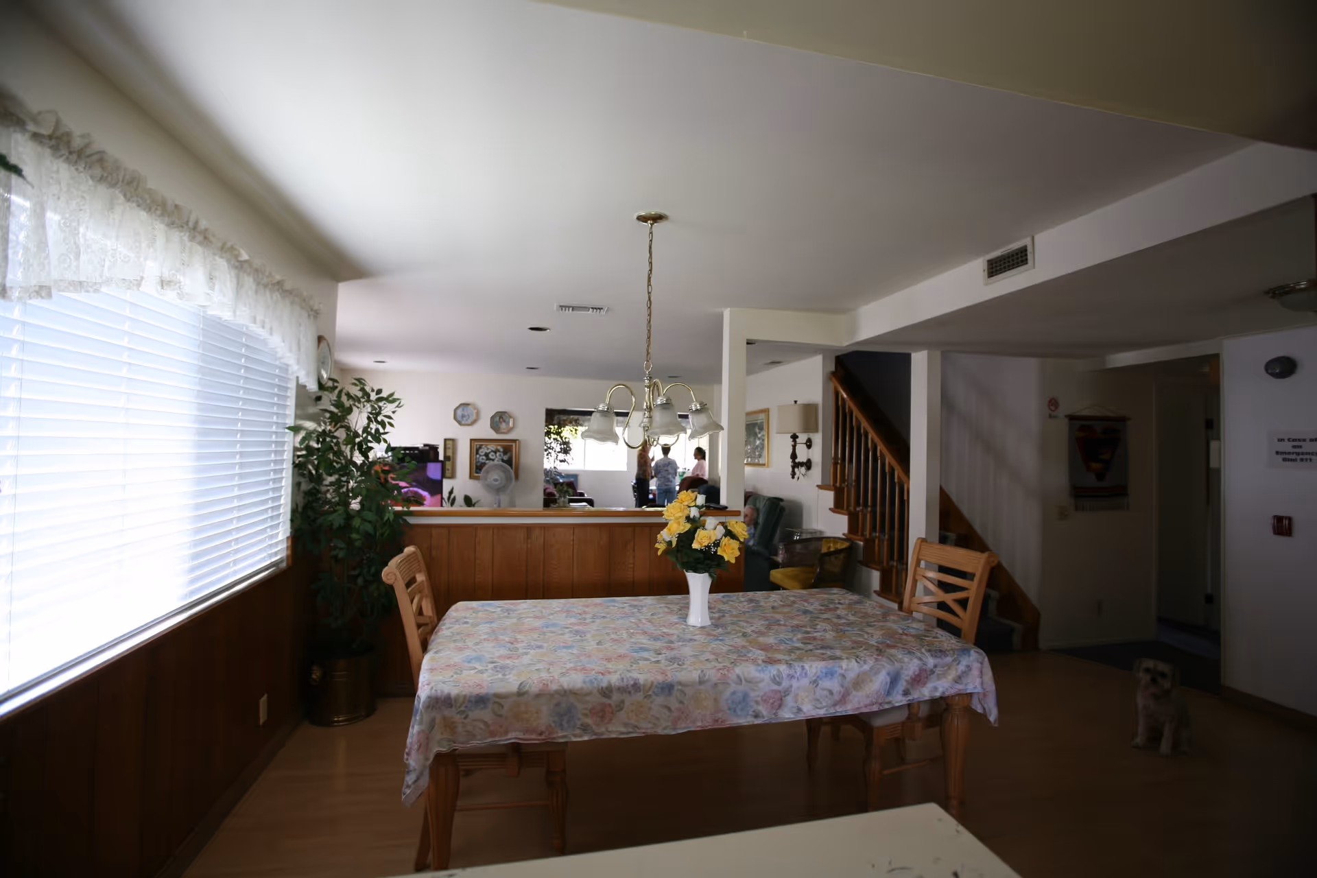 Interior view of a senior living facility dining area with a wooden table covered by a floral tablecloth and a vase of yellow flowers on top. Two wooden chairs are placed at the table. Large window with white blinds and lace valance on the left side. In the background, there is a living room area with people standing and sitting, a staircase with wooden railing, and a small dog sitting on the floor near the entrance.