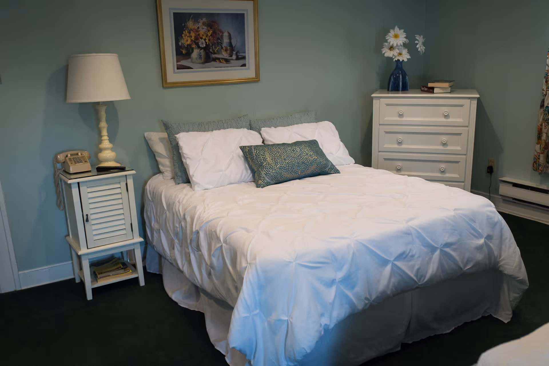 A neatly made bed with white bedding and multiple pillows in a room with light green walls. To the left of the bed is a small white nightstand with a vintage telephone and a lamp. On the right side of the bed is a white chest of drawers with a blue vase holding white flowers and a couple of books on top. A framed floral painting hangs on the wall above the bed.
