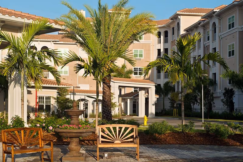 Outdoor courtyard area of Diamond Oaks Village featuring palm trees, a stone fountain, wooden benches, and a multi-story building with beige walls and red-tiled roofs in the background.
