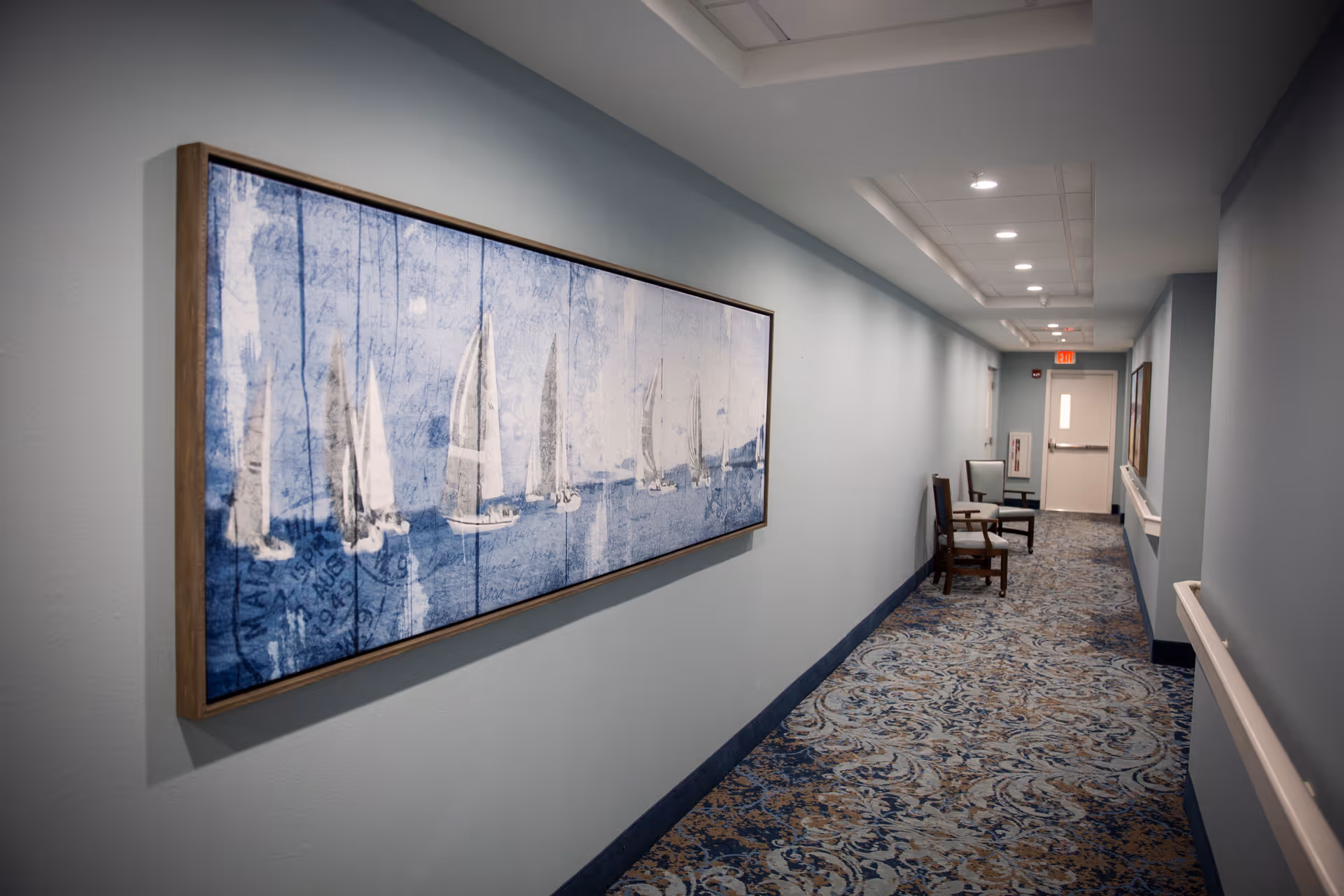 A long hallway with light blue walls and patterned carpet. On the left wall, there is a large framed artwork depicting sailboats on water in shades of blue. At the end of the hallway, there are two chairs placed against the right wall near a door with an exit sign above it. The ceiling has recessed lighting.