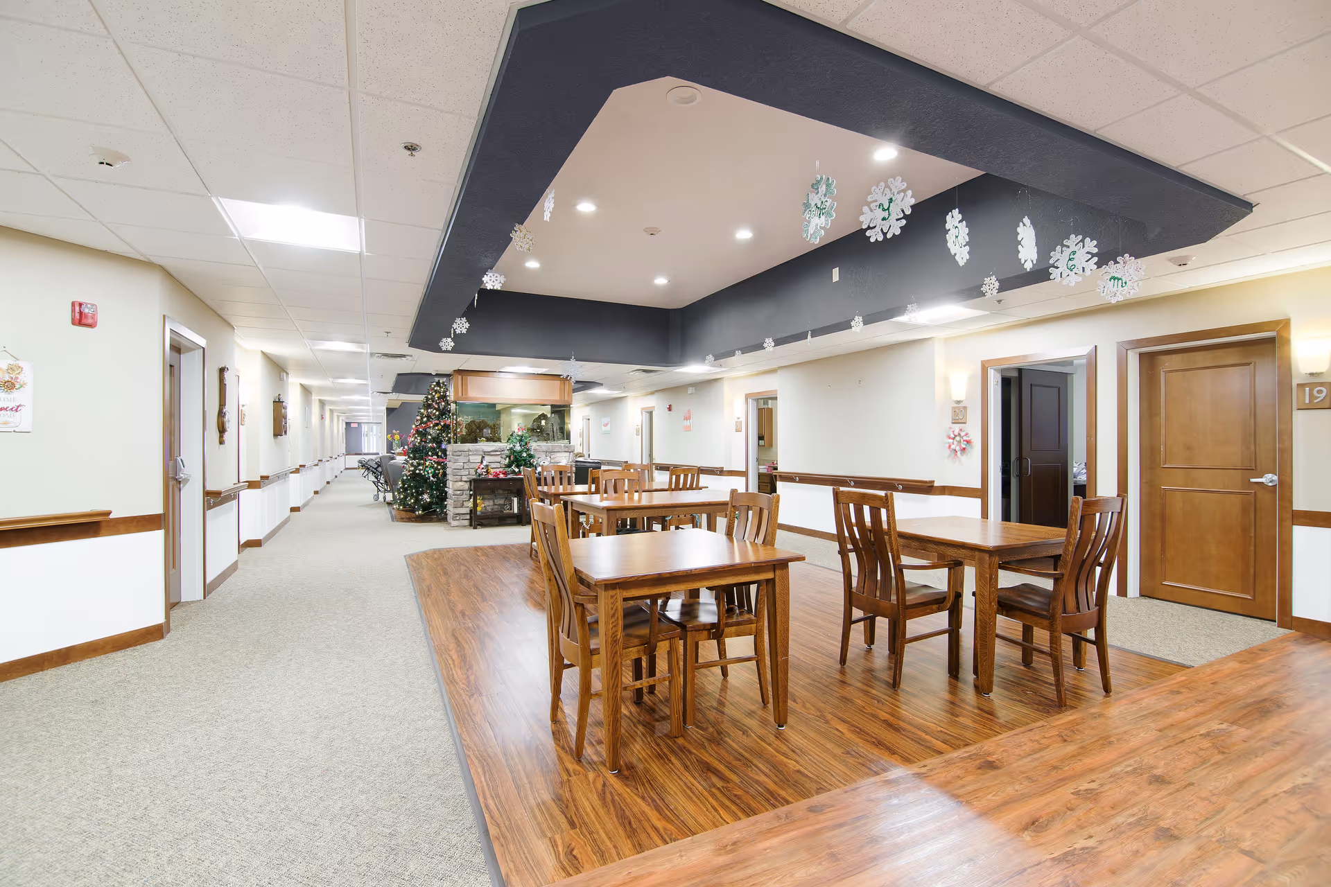 Interior view of a senior living facility hallway with a seating area featuring wooden tables and chairs on a wood floor section. The hallway has beige carpet, cream-colored walls with wooden trim, and several doors along the corridor. There are Christmas decorations including a decorated Christmas tree and hanging snowflakes from the ceiling.