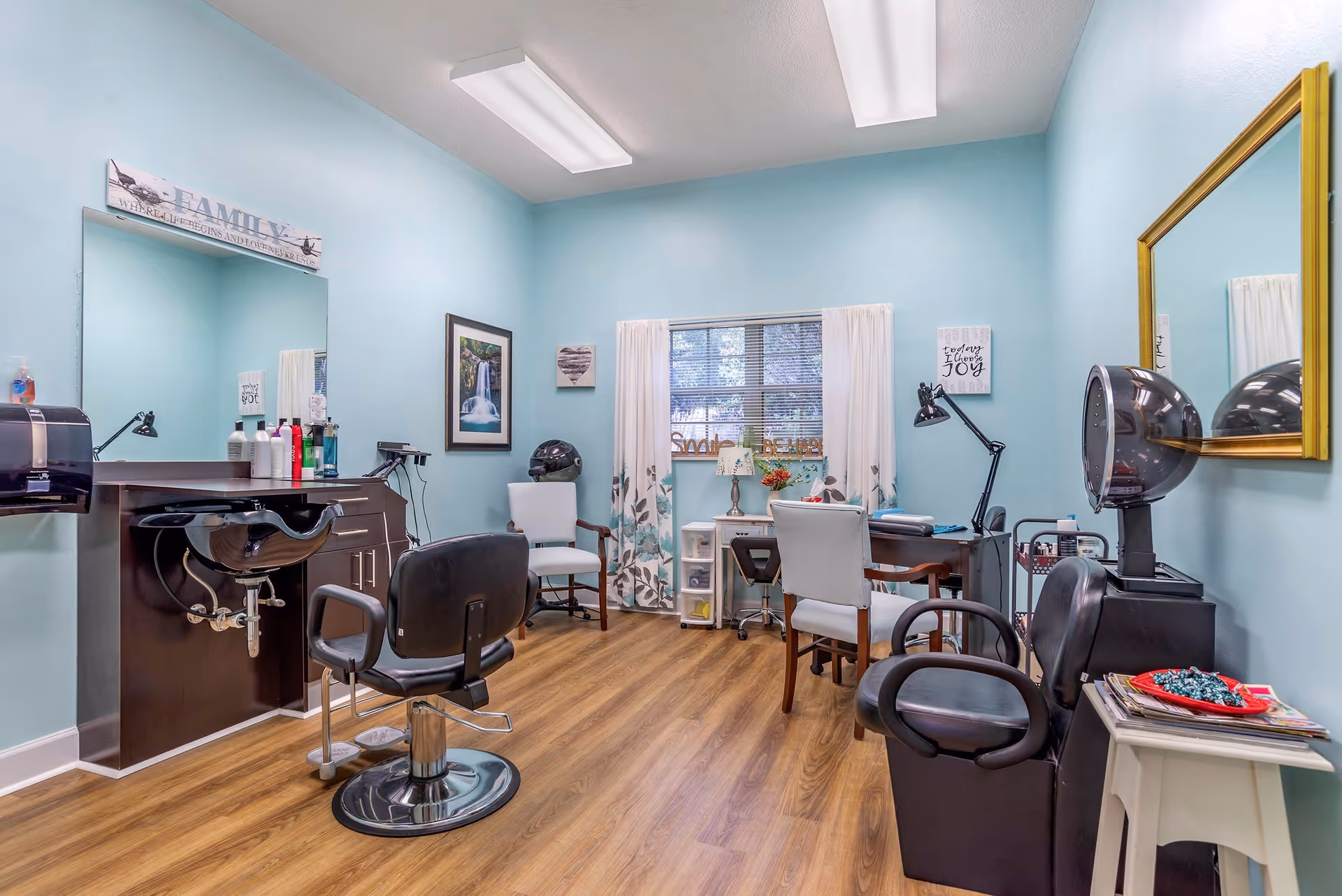 A bright and clean salon room with light blue walls and wood flooring. The room features a black salon chair in front of a sink and mirror, a hair dryer chair with a hooded dryer, two white chairs, a small desk with a lamp and flowers, and decorative wall art including a framed waterfall picture and inspirational signs. A window with white curtains lets in natural light.