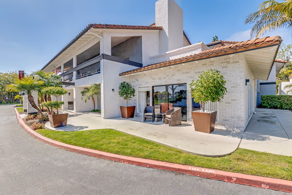 Exterior view of a two-story senior living facility building with white walls and a red tile roof. There are potted plants and outdoor seating on the patio area. The sky is clear and blue.