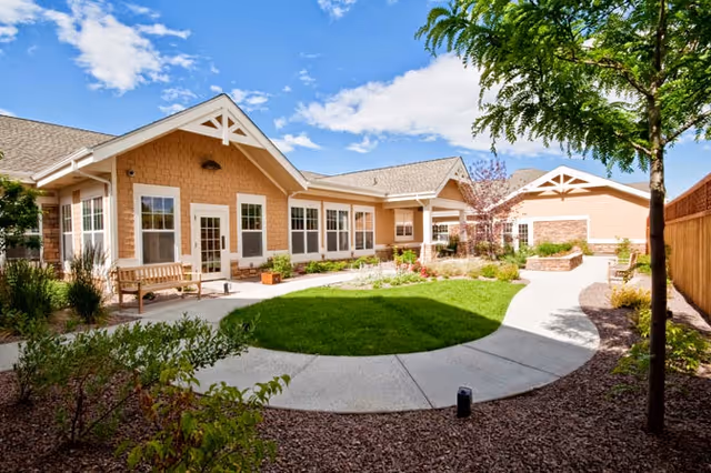 Sunny courtyard with a curved concrete path, lawn, benches, and a single-story beige assisted-living building.
