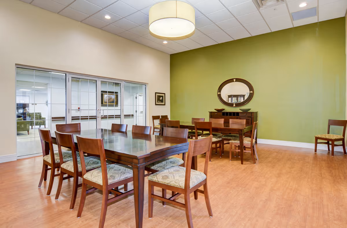 A dining room with two wooden tables and multiple wooden chairs with patterned cushions. The room has light-colored walls with one green accent wall, a round mirror above a wooden sideboard, and a large circular ceiling light. There are glass sliding doors leading to another room with more seating visible.