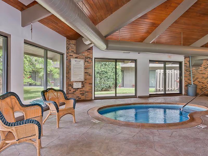 Indoor hot tub area with a round hot tub surrounded by a tiled floor. There are two wicker chairs with cushions placed near large windows that look out onto a green outdoor area. The ceiling has wooden panels and exposed ductwork.
