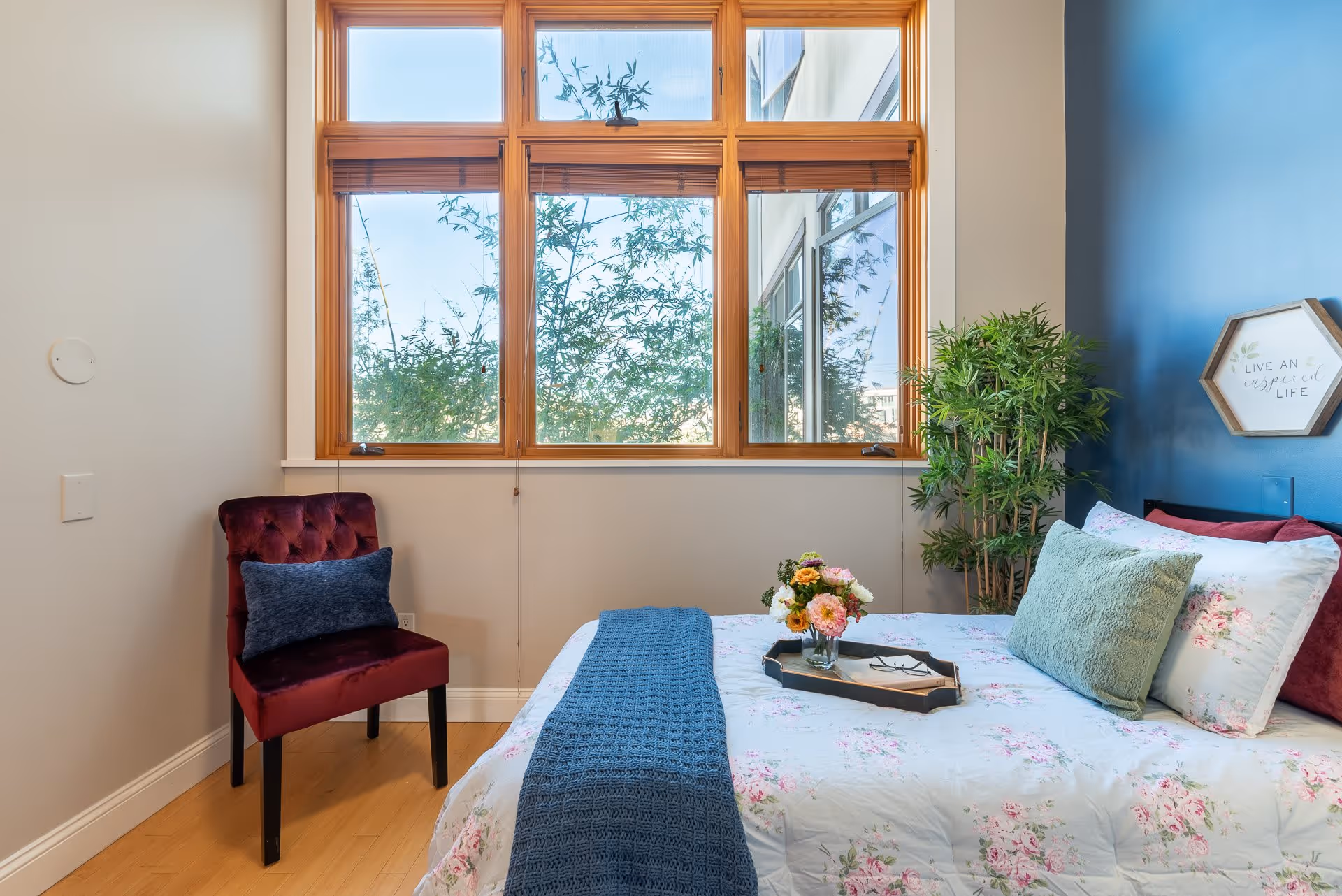 A cozy bedroom with a large window featuring wooden frames and a view of greenery outside. The bed is made with floral bedding, a blue knitted throw, and several pillows. A tray with flowers and reading glasses rests on the bed. To the left, there is a burgundy upholstered chair with a blue cushion. A green potted plant is placed near the bed against a blue accent wall with a hexagonal wall decoration that reads 'Live an inspired life.'