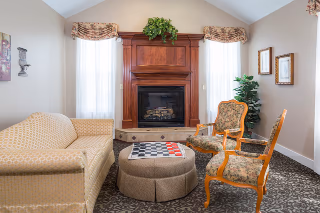 A cozy living room with a beige patterned sofa, two floral upholstered wooden armchairs, and a round ottoman with a checkerboard on top. The room features a wooden fireplace with a plant on the mantel, two tall windows with sheer white curtains and floral valances, a potted plant in the corner, and framed artwork on the walls.