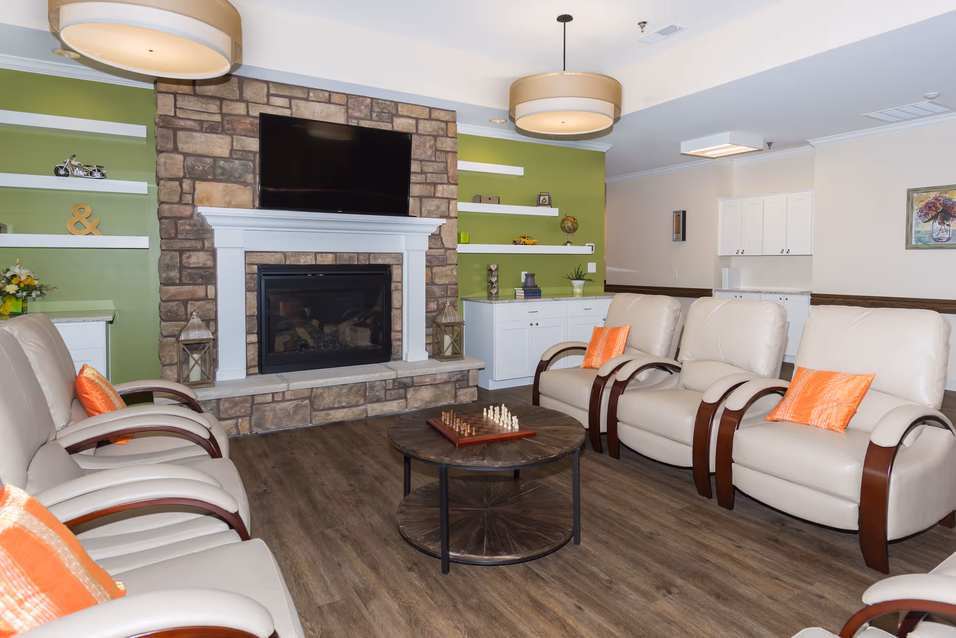 A cozy living room area with beige leather armchairs arranged around a round wooden coffee table with a chess set on it. The room features a stone fireplace with a mounted flat-screen TV above it, green accent walls with white shelves displaying decorative items, and white cabinetry. The floor is wood, and there are two large ceiling lights.