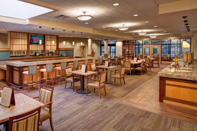 A spacious dining area in a senior living facility featuring multiple wooden tables and chairs arranged neatly. There is a long counter with bar stools on one side and a buffet area with granite countertops on the other. The room is well-lit with ceiling lights and has large windows and glass doors at the far end, allowing natural light to enter. The decor includes warm wood tones and a carpeted floor with a wood-patterned section near the buffet.