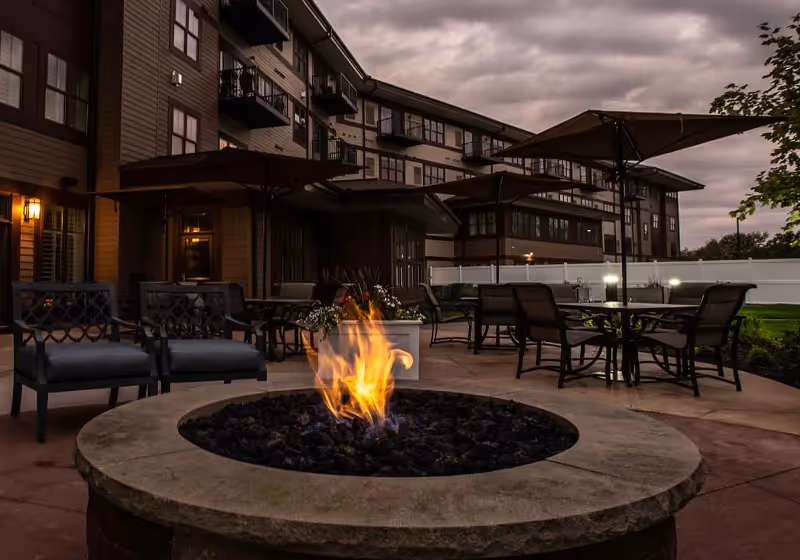 Outdoor patio area at dusk with a round fire pit in the foreground surrounded by chairs. Several tables with umbrellas and chairs are arranged on the patio. A multi-story building with balconies and lit windows is visible in the background under a cloudy sky.