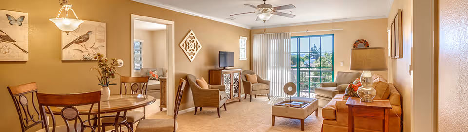 A bright and cozy living room and dining area in a senior living facility. The dining area has a round wooden table with four chairs and a hanging light fixture above. The living room features a beige sofa, two armchairs, a wooden coffee table, a side table with a lamp, and a TV on a wooden cabinet. Large sliding glass doors with vertical blinds open to a balcony with a view of trees and a clear sky. The walls are painted a warm beige color and decorated with framed artwork and a decorative wall piece.