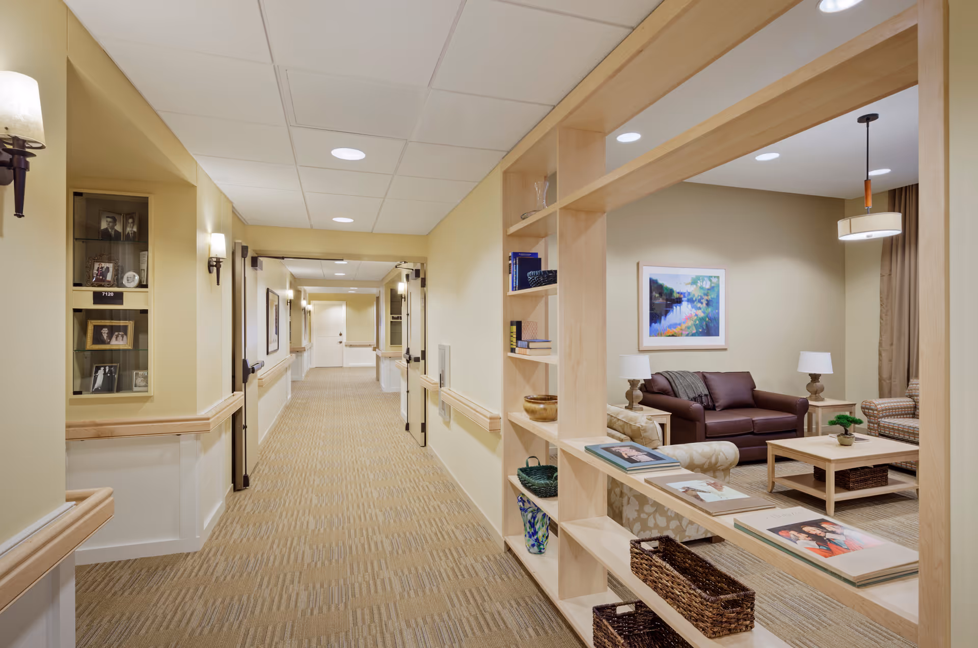A well-lit hallway in a senior living facility with beige walls and carpeted floor. On the right side, there is a built-in wooden shelving unit displaying books, baskets, and decorative items. Beyond the shelving unit is a cozy seating area with a brown leather sofa, an armchair, a wooden coffee table, two table lamps, and a colorful framed painting on the wall. The hallway has wall-mounted lights and handrails along both sides.
