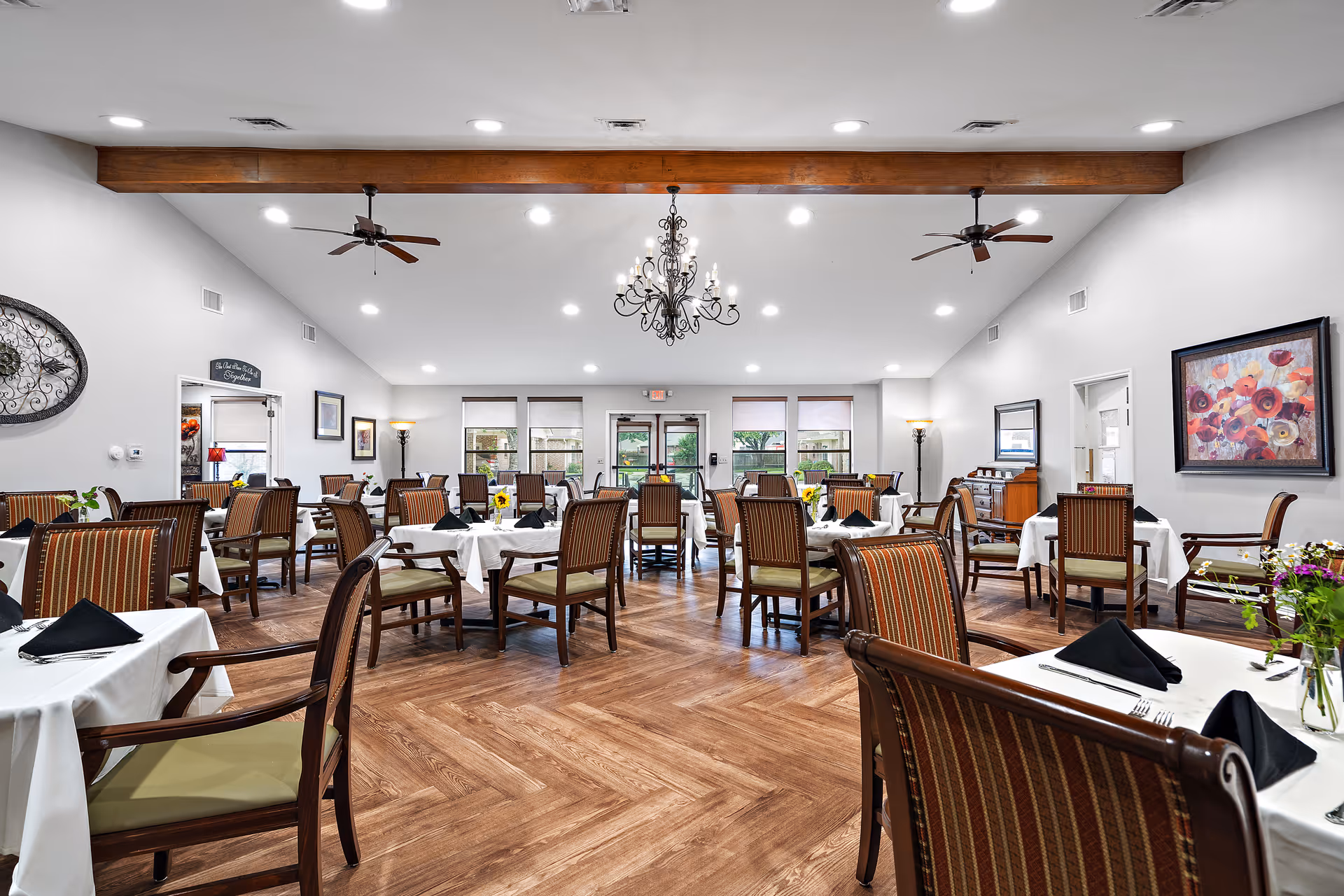 A spacious dining room with multiple tables covered in white tablecloths, each set with black napkins and silverware. The room features wooden chairs with striped upholstery, hardwood flooring, ceiling fans, and a decorative chandelier. Large windows and glass doors at the far end let in natural light, and the walls are adorned with framed artwork and decorative pieces.