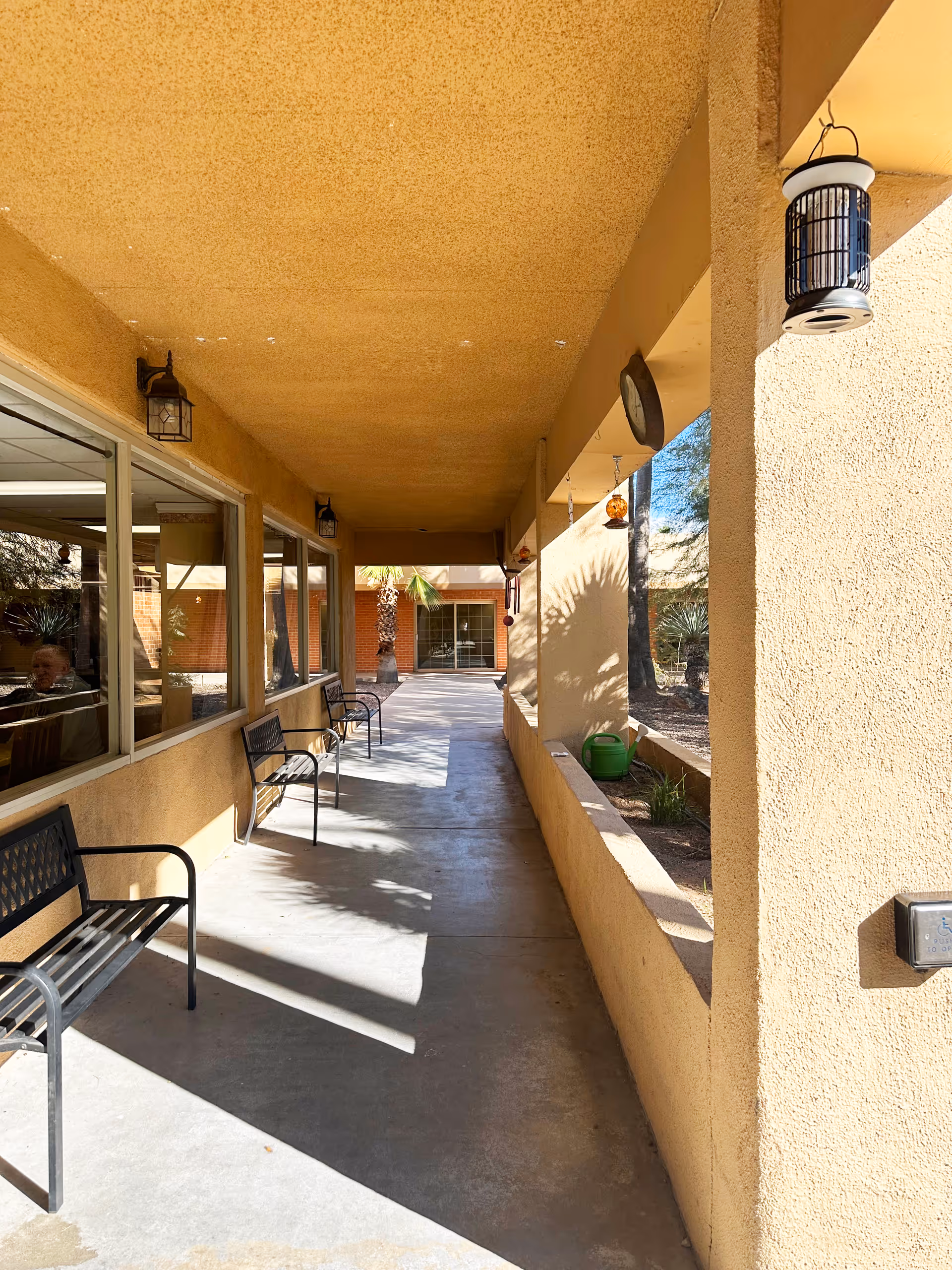Covered outdoor walkway with benches along windows, hanging lanterns, and a view into a courtyard with a palm tree.