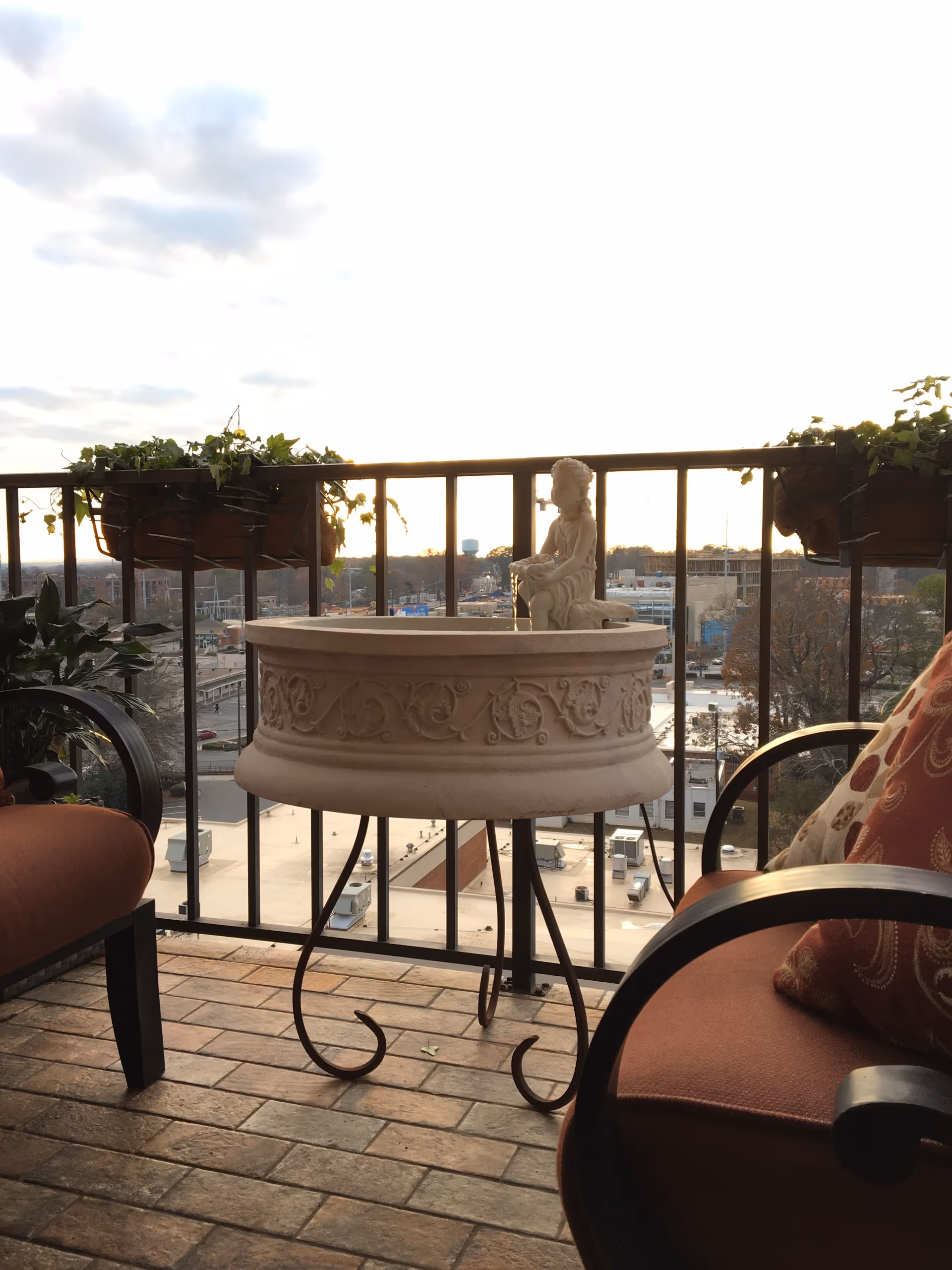 A balcony with two cushioned chairs on either side, a decorative round stone planter with a small statue on a metal stand in the center, and potted plants hanging on the railing overlooking a cityscape in the background.