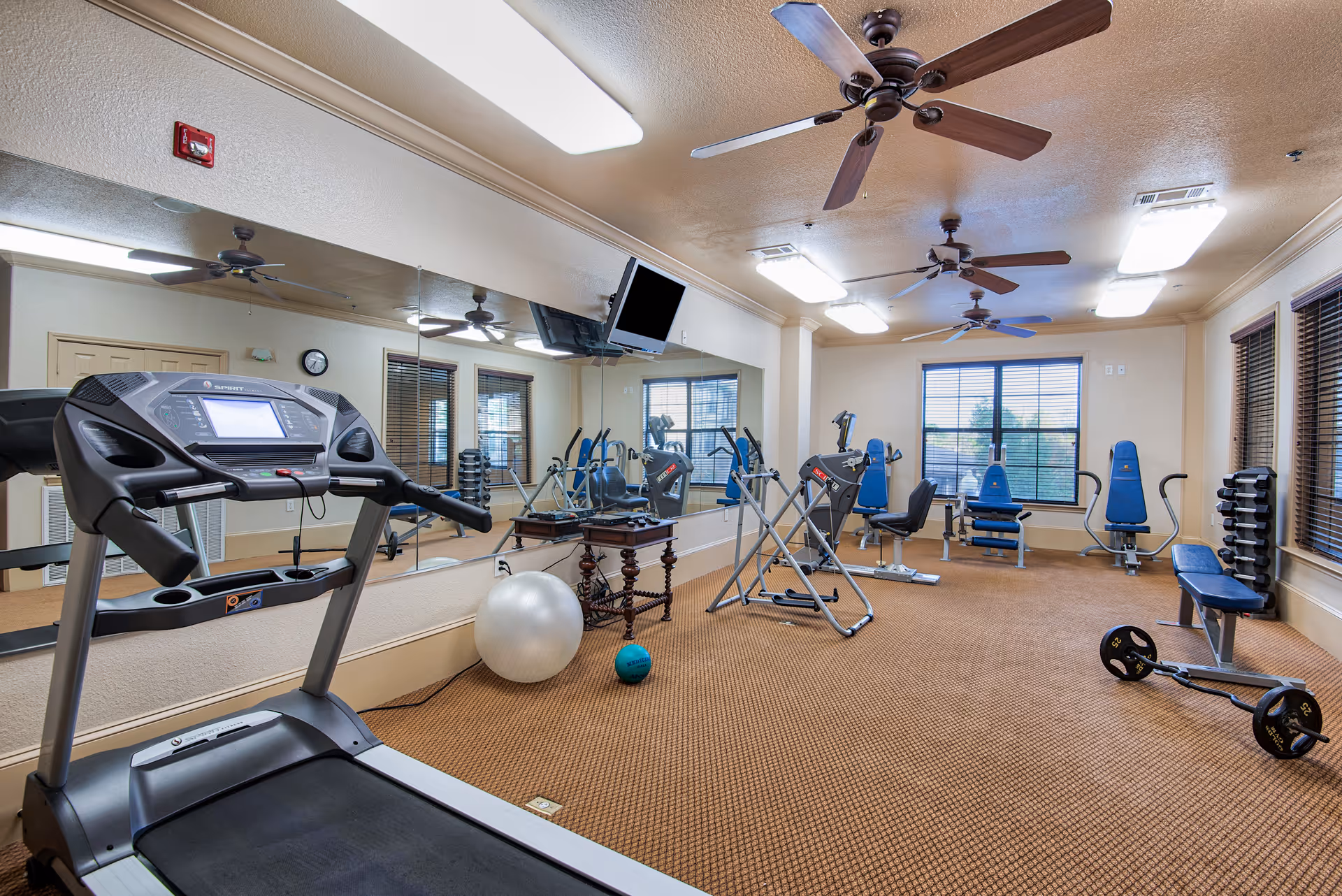 A fitness room with various exercise equipment including a treadmill, weight bench, dumbbells, exercise balls, and resistance machines. The room has large windows with blinds, ceiling fans, and a wall covered with mirrors.