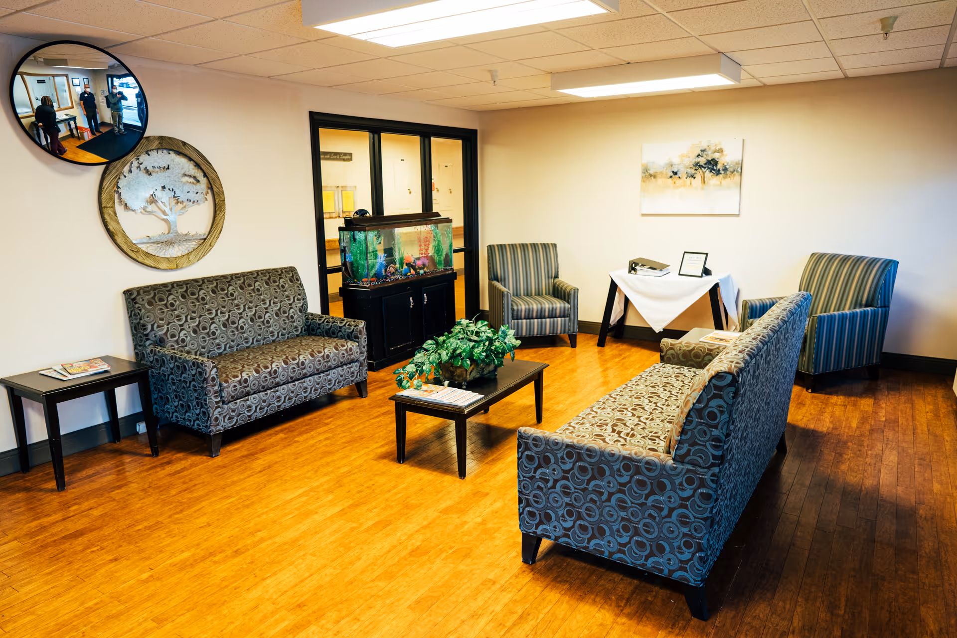 A cozy seating area in a senior living facility with two patterned sofas, two striped armchairs, a coffee table with a plant, a side table with magazines, and an aquarium on a black stand. The room has wooden flooring, beige walls, a painting on the wall, and a convex security mirror showing three people in the hallway.