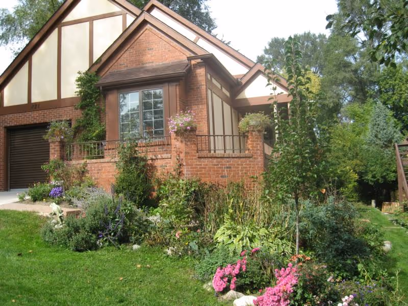 Exterior view of a brick and wood-paneled assisted living facility building with a sloped roof, surrounded by a well-maintained garden with various green plants and colorful flowers.