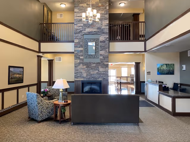 Spacious two-story senior living lobby with a central stone fireplace, seating area, chandelier, and reception desk.