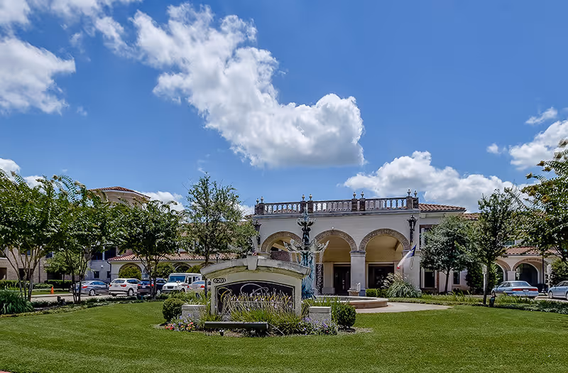 Exterior view of Conservatory At Alden Bridge senior living facility with a well-maintained green lawn, trees, a decorative fountain, and a clear blue sky with some clouds.