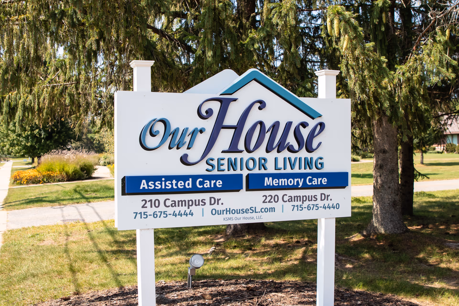 Outdoor sign for "Our House Senior Living" showing "Assisted Care" and "Memory Care" on a lawn with trees in the background.