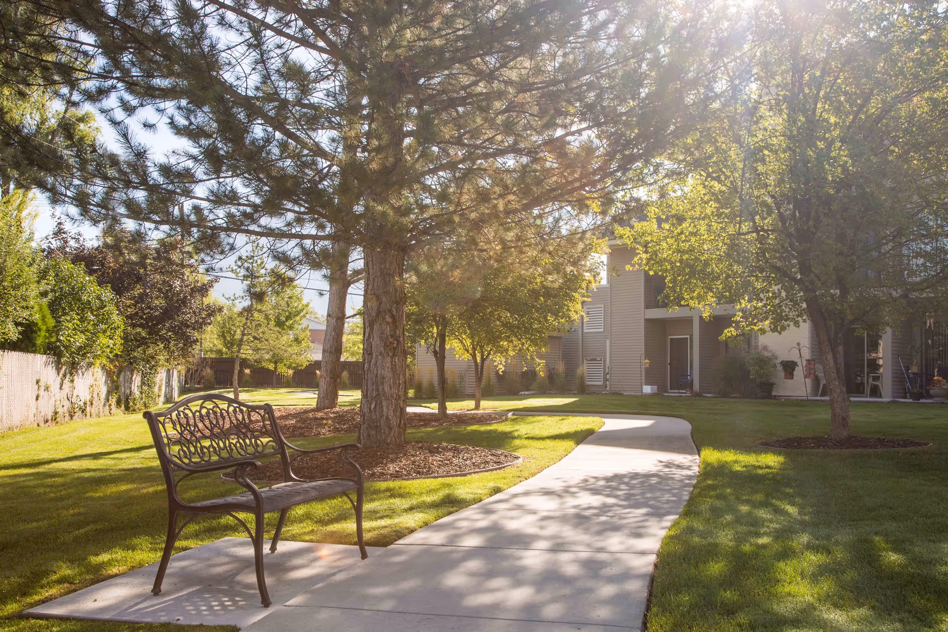 Sunlit courtyard with a metal bench beside a winding concrete path, trees, grass, and apartment building in the background.