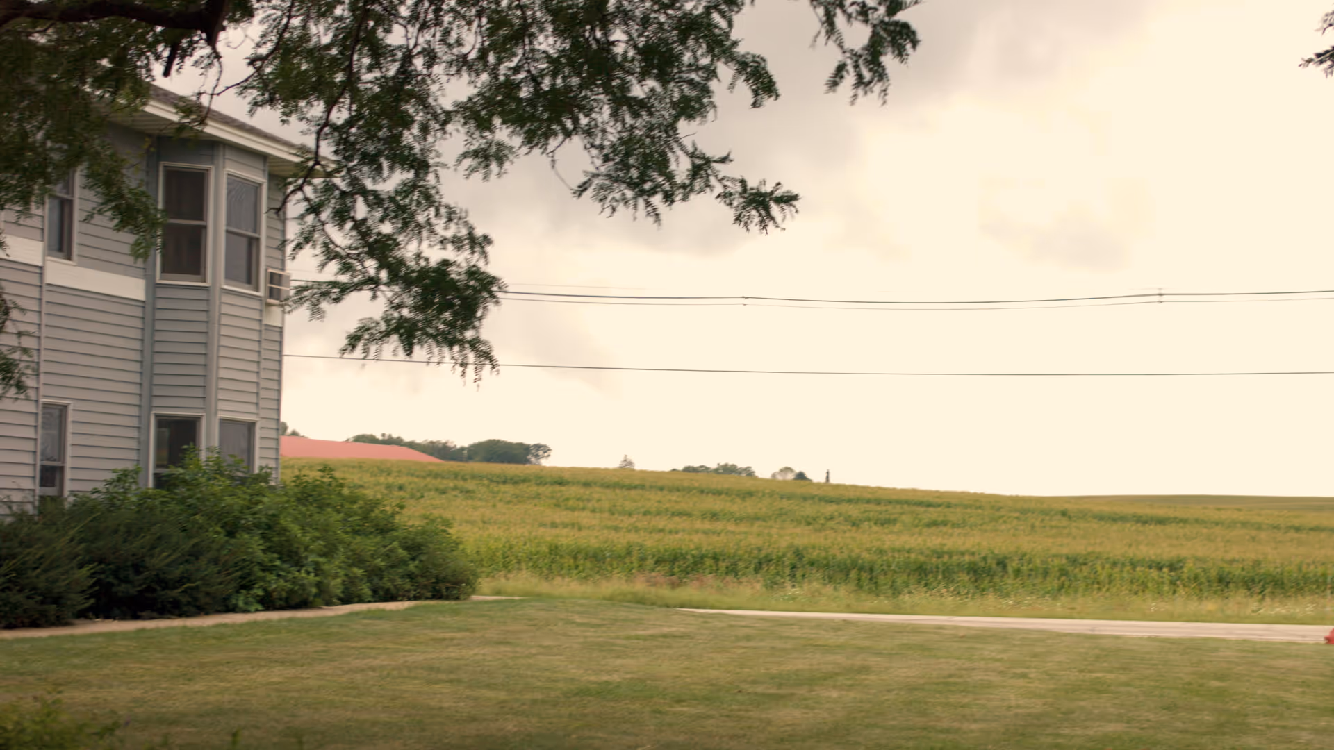 Side view of a gray two-story building with several windows, surrounded by green bushes and a large grassy lawn. In the background, there is a wide field of crops under a cloudy sky, with some trees and a red-roofed structure visible in the distance.