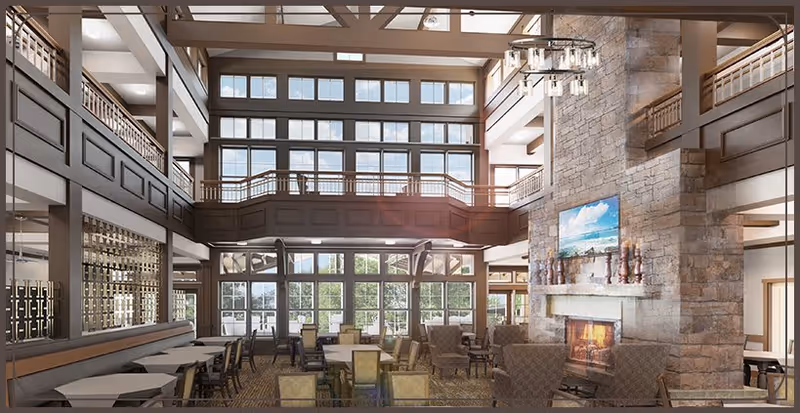 Spacious and well-lit living room area with high ceilings and large windows allowing natural light to fill the space. The room features multiple seating arrangements with chairs and tables, a large stone fireplace with a mounted TV above it, and a second-floor balcony overlooking the area.