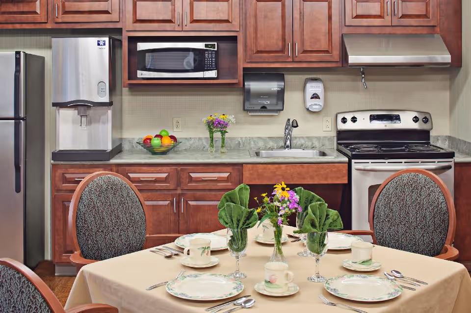 A kitchen area with wooden cabinets, a stainless steel refrigerator, a microwave, a water dispenser, a sink, and a stove. In the foreground, there is a dining table set with plates, cups, utensils, green folded napkins in glasses, and a small vase with flowers. The chairs around the table have patterned upholstery.
