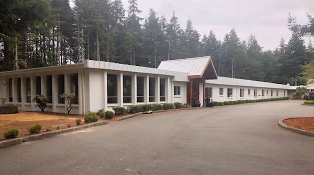 Exterior view of a single-story building with large windows and a peaked entrance, surrounded by trees and landscaping with bushes and mulch beds.