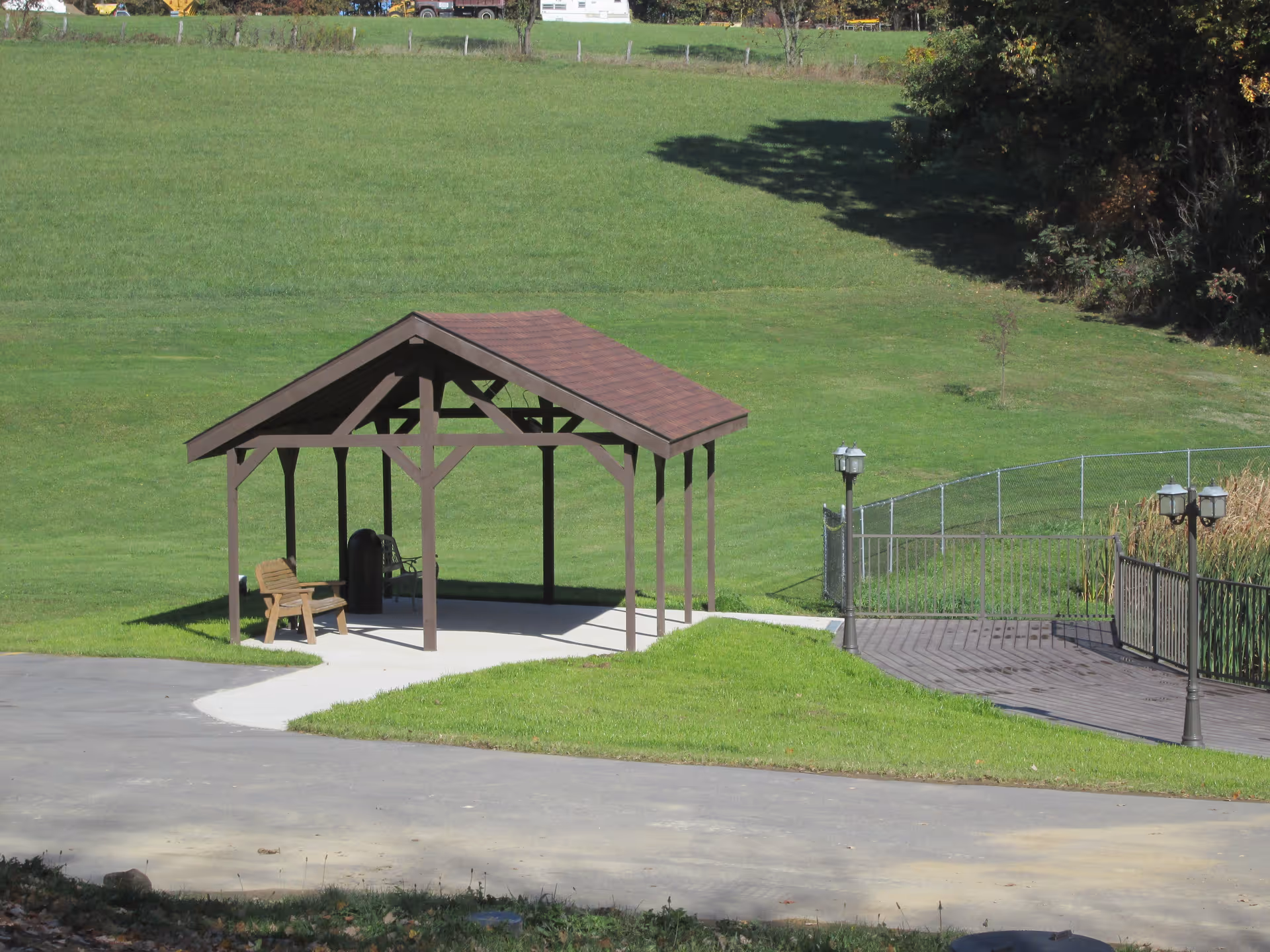 Wooden gazebo with a bench on a concrete pad set in a grassy lawn near a fenced boardwalk and lampposts.