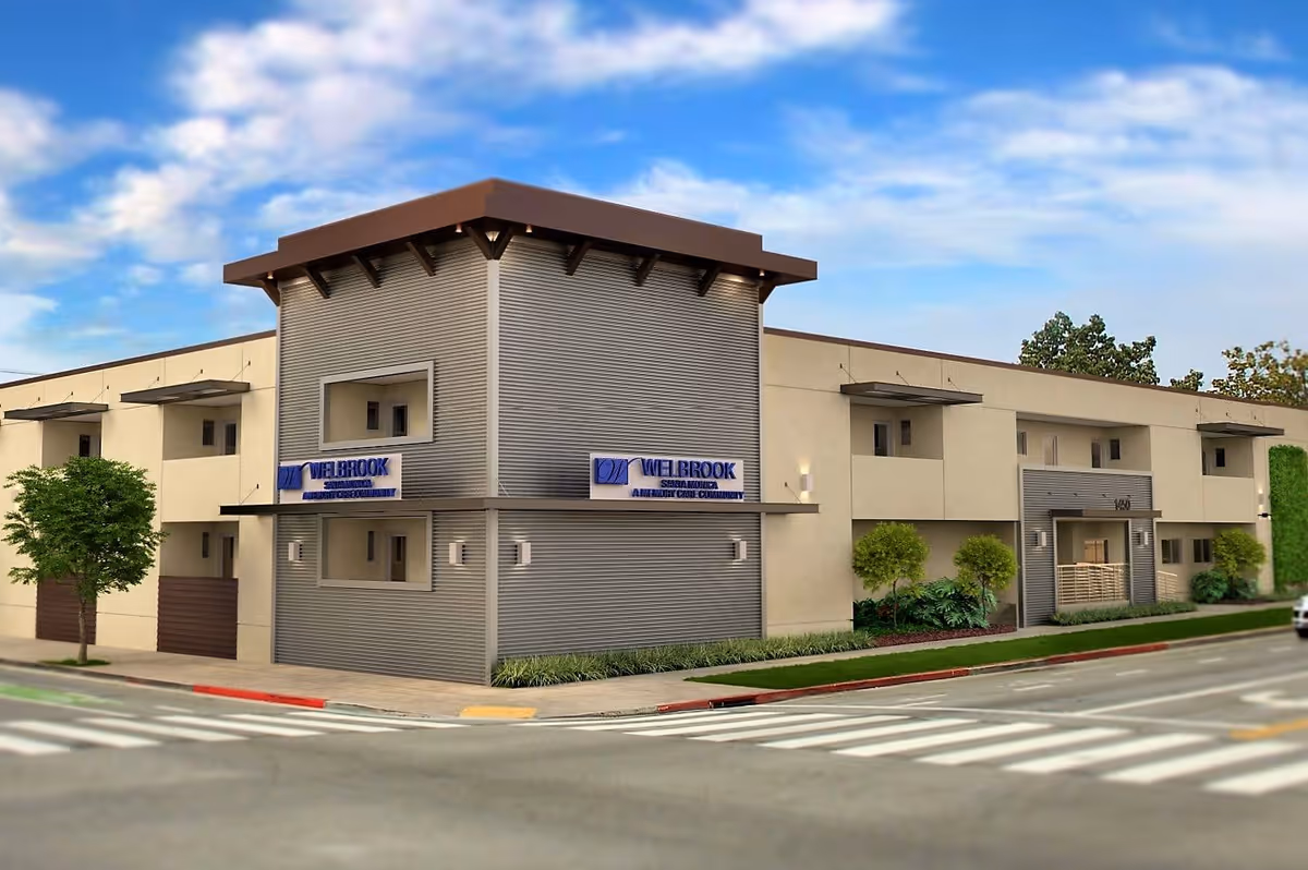 Exterior view of Welbrook Santa Monica, a modern two-story assisted living community building with beige and gray walls, large windows, and a corner entrance. The building is situated at a street intersection with crosswalks, trees, and a clear blue sky with some clouds.