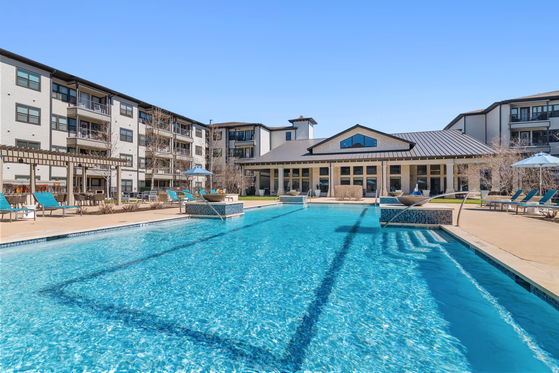 Outdoor swimming pool with lounge chairs and umbrellas in front of a multi-story senior living building under a clear blue sky.