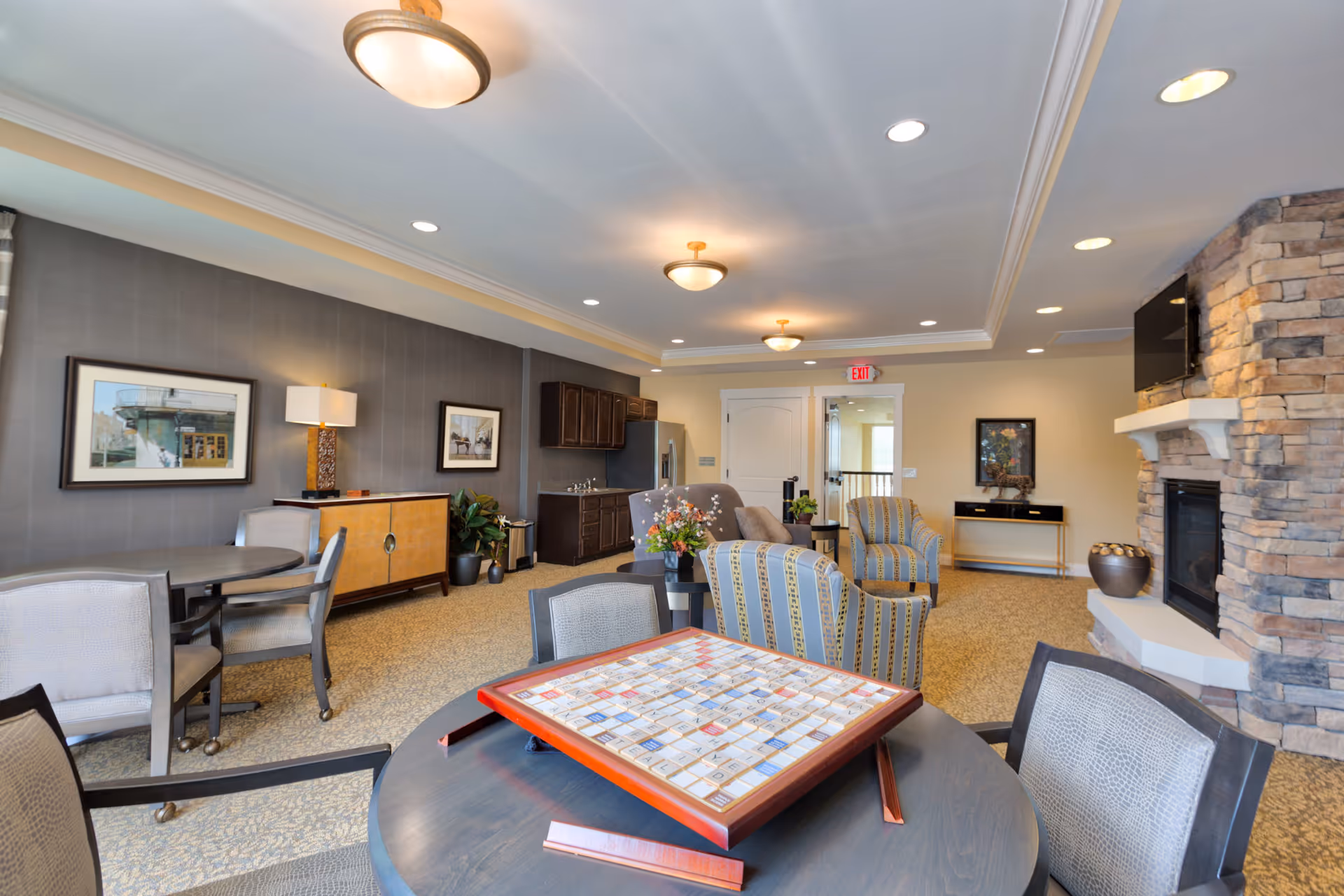 A well-lit common lounge area with a Scrabble board on a round table in the foreground. The room features comfortable armchairs, a stone fireplace with a mounted TV, framed artwork on the walls, and a small kitchenette with dark cabinets and a refrigerator. The carpeted floor and ceiling lights create a cozy atmosphere.