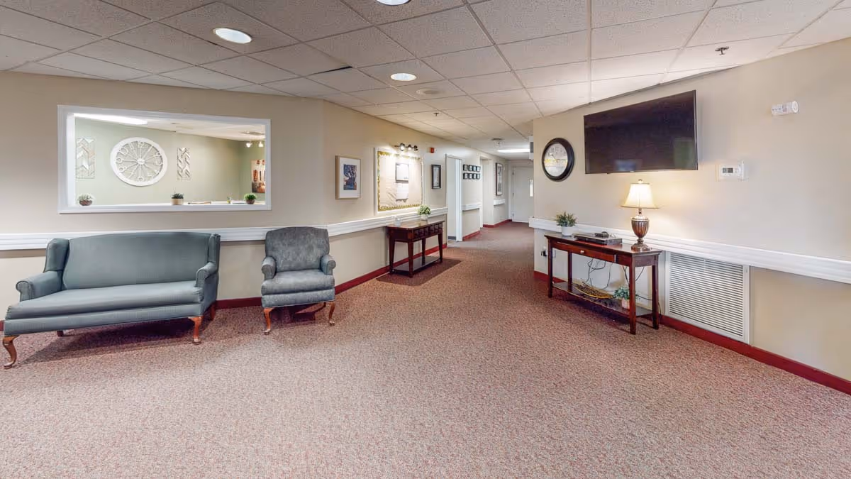 Interior hallway of a senior living facility with carpeted floor, beige walls, and ceiling tiles. There are two upholstered chairs on the left side under a large window opening to another room. On the right side, a wooden console table holds a lamp and a flat-screen TV mounted on the wall above it. Further down the hallway are framed pictures and a bulletin board with lighting above it.