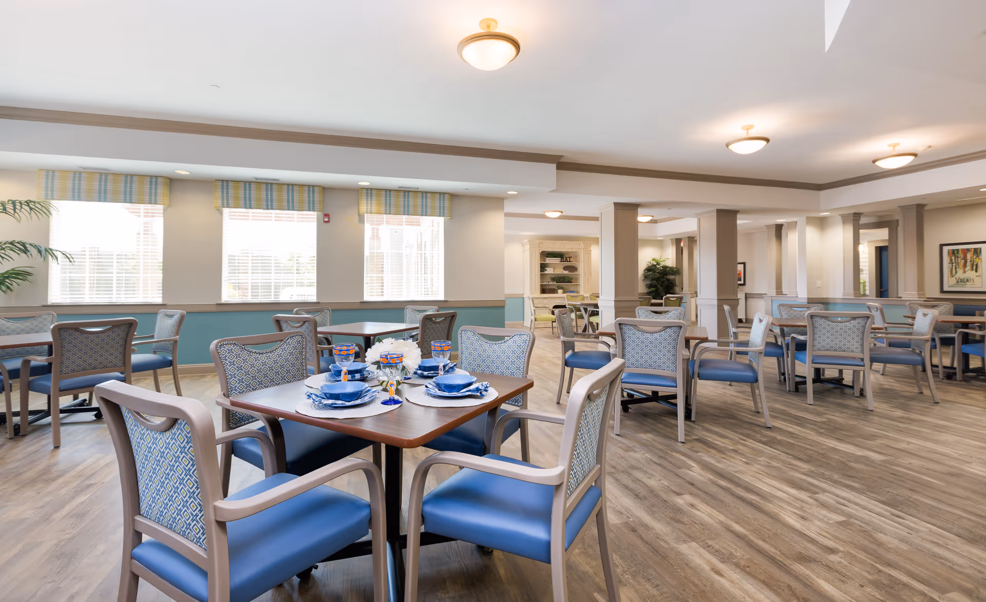 A bright and spacious dining room in a senior living facility with multiple tables and chairs arranged neatly. The tables are set with blue plates, bowls, glasses, and napkins, and a small floral centerpiece is on the closest table. Large windows with striped valances allow natural light to fill the room, which features wood flooring and neutral-colored walls with blue wainscoting.