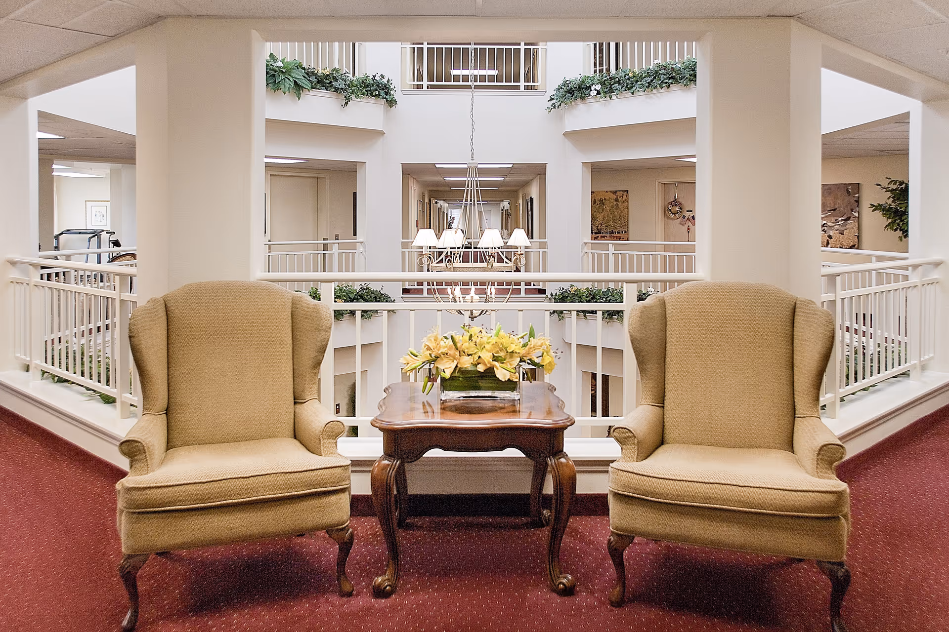 Two beige upholstered armchairs with wooden legs are positioned on either side of a wooden table with a floral arrangement in a bright indoor hallway area with white railings and red carpet. The space overlooks an open atrium with plants and a hanging chandelier.