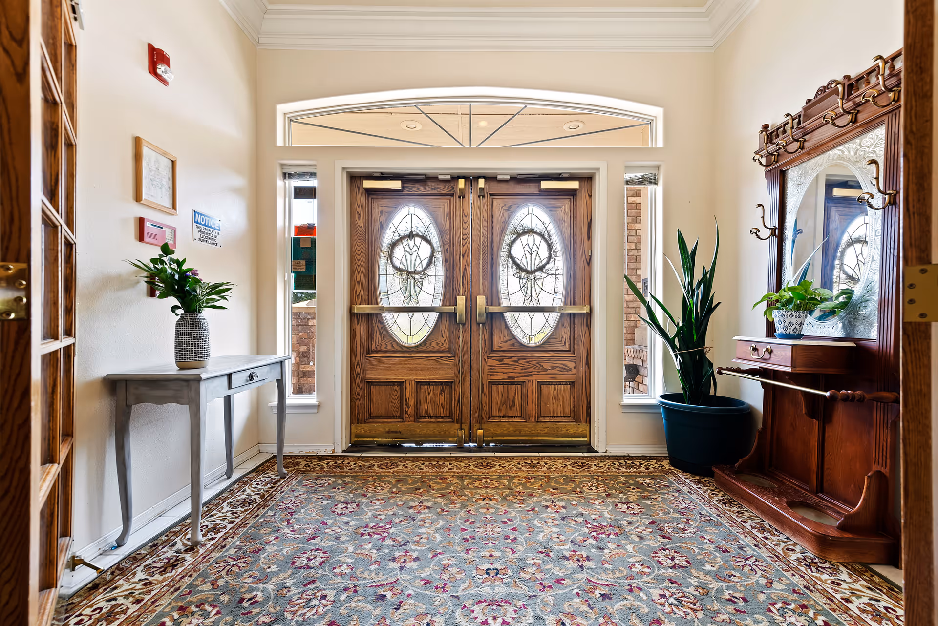 Entrance area of Autumn Wind Assisted Living featuring double wooden doors with decorative glass panels, a patterned rug on the floor, a small table with a potted plant on the left, and a wooden coat rack with a mirror and plants on the right.