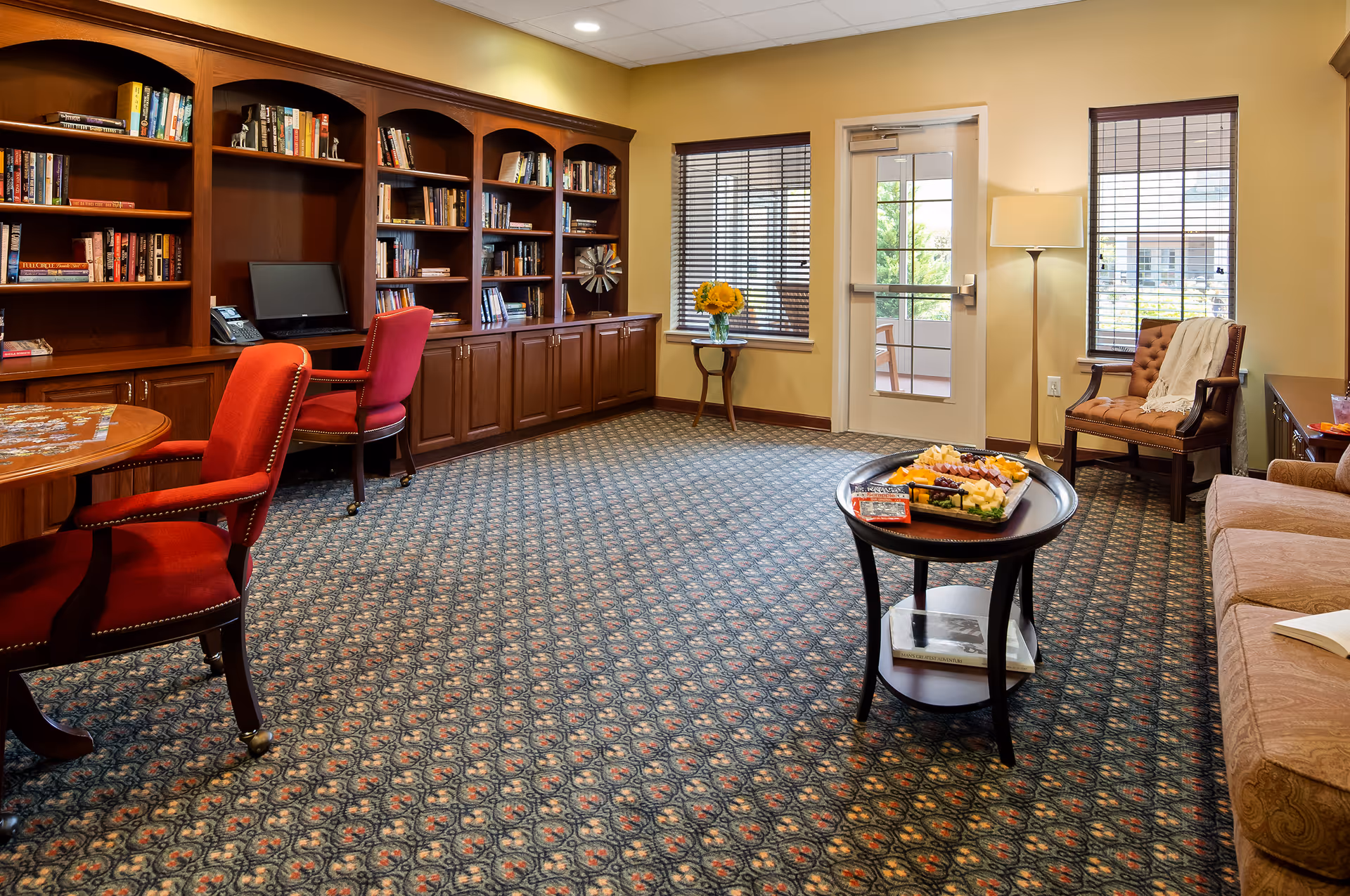 A cozy common room with patterned carpet, wooden bookshelves filled with books, a computer on a desk, red upholstered chairs, a round table with a puzzle, a small round table with a tray of snacks, a beige sofa, an armchair with a white throw, a floor lamp, two windows with blinds, and a glass door leading outside.