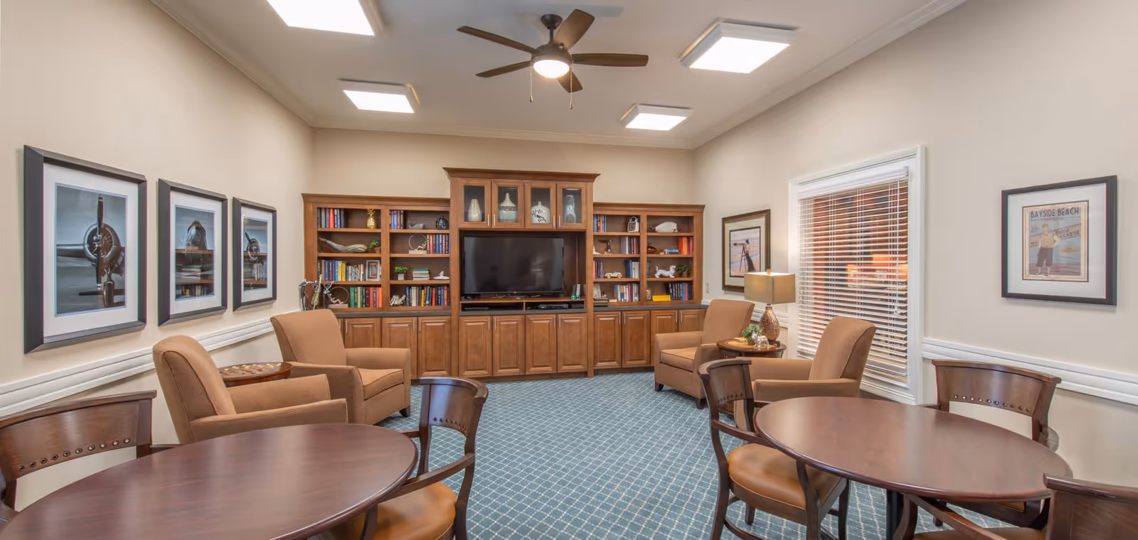 A cozy common area with a blue patterned carpet, beige walls, and a ceiling fan. The room features a large wooden entertainment center with a TV and bookshelves filled with books and decorative items. There are four beige armchairs arranged around a small wooden table, and two round wooden tables with chairs. The walls are decorated with framed pictures, and a window with white blinds allows natural light into the room.