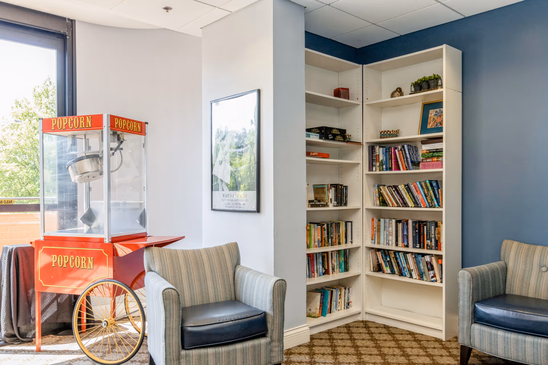 A cozy corner of a room featuring a vintage-style red popcorn machine on wheels, two striped armchairs with dark cushions, and a white corner bookshelf filled with books and small decorative items. The walls are painted light gray and blue, and there is a framed picture hanging on the wall between the popcorn machine and the bookshelf.