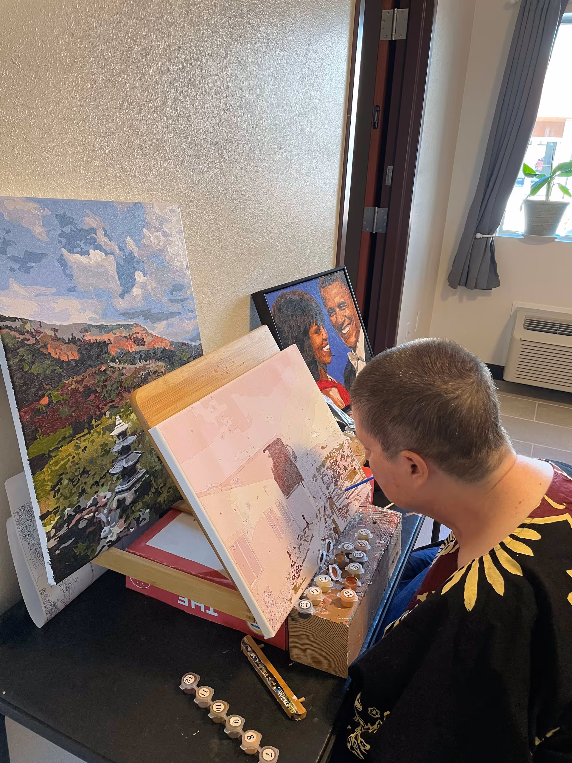A person with short hair is seated at a table, working on a paint-by-numbers canvas. Several completed paintings are propped up nearby, including a landscape and a portrait of a smiling couple. The room has a window with a potted plant and an air conditioning unit beneath it.