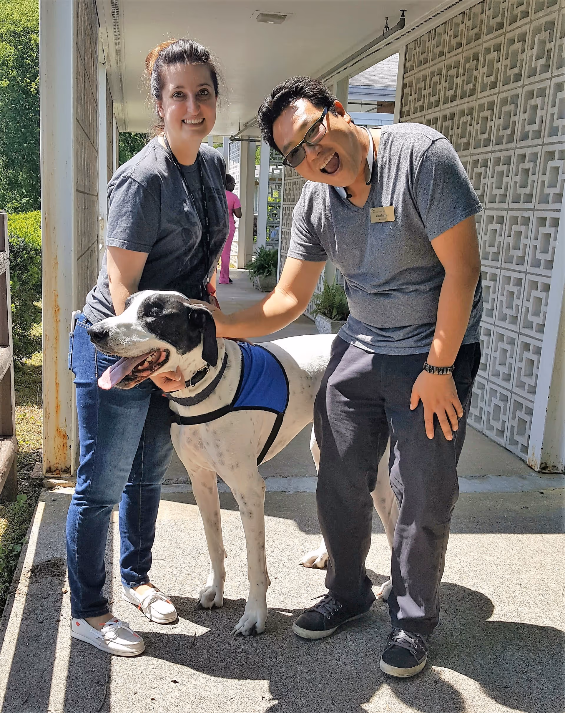 Two staff members smile while petting a large dog in a well-lit outdoor area of the facility.