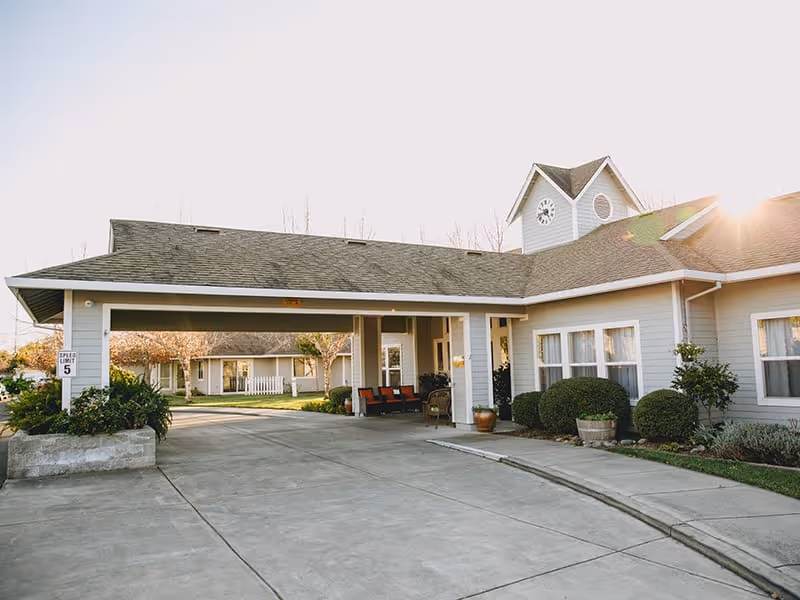 Exterior view of Sonoma Hills Retirement Living Community showing a covered driveway entrance with seating, surrounded by well-maintained bushes and trees under a clear sky with sunlight.