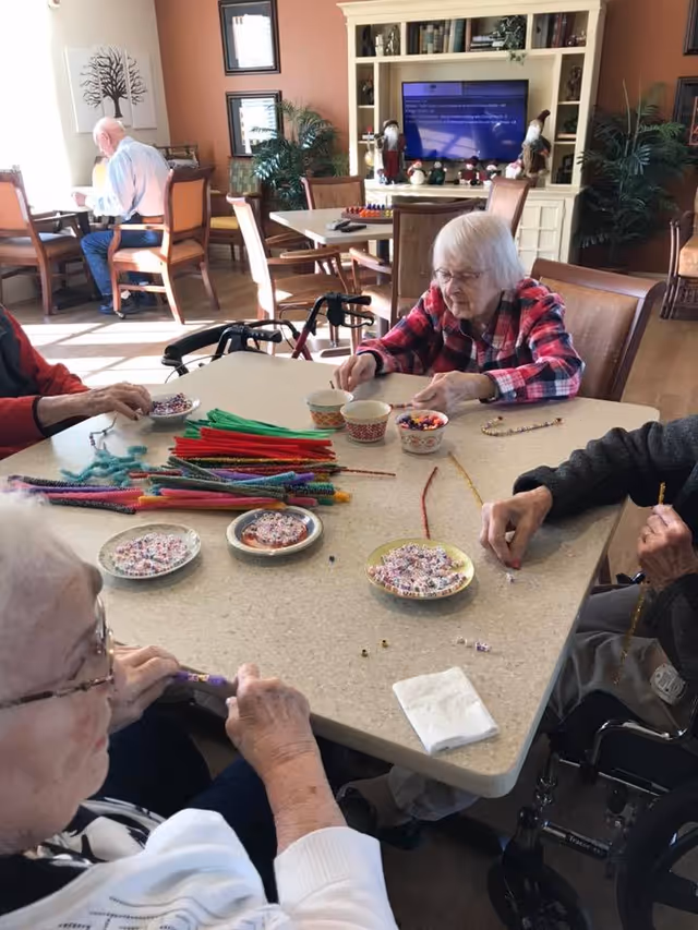 Several elderly individuals seated around a table in a well-lit room, engaging in a craft activity with colorful pipe cleaners and beads. The room has wooden chairs, a large white cabinet with a TV, and decorative plants. Another elderly person is seen sitting at a separate table in the background.