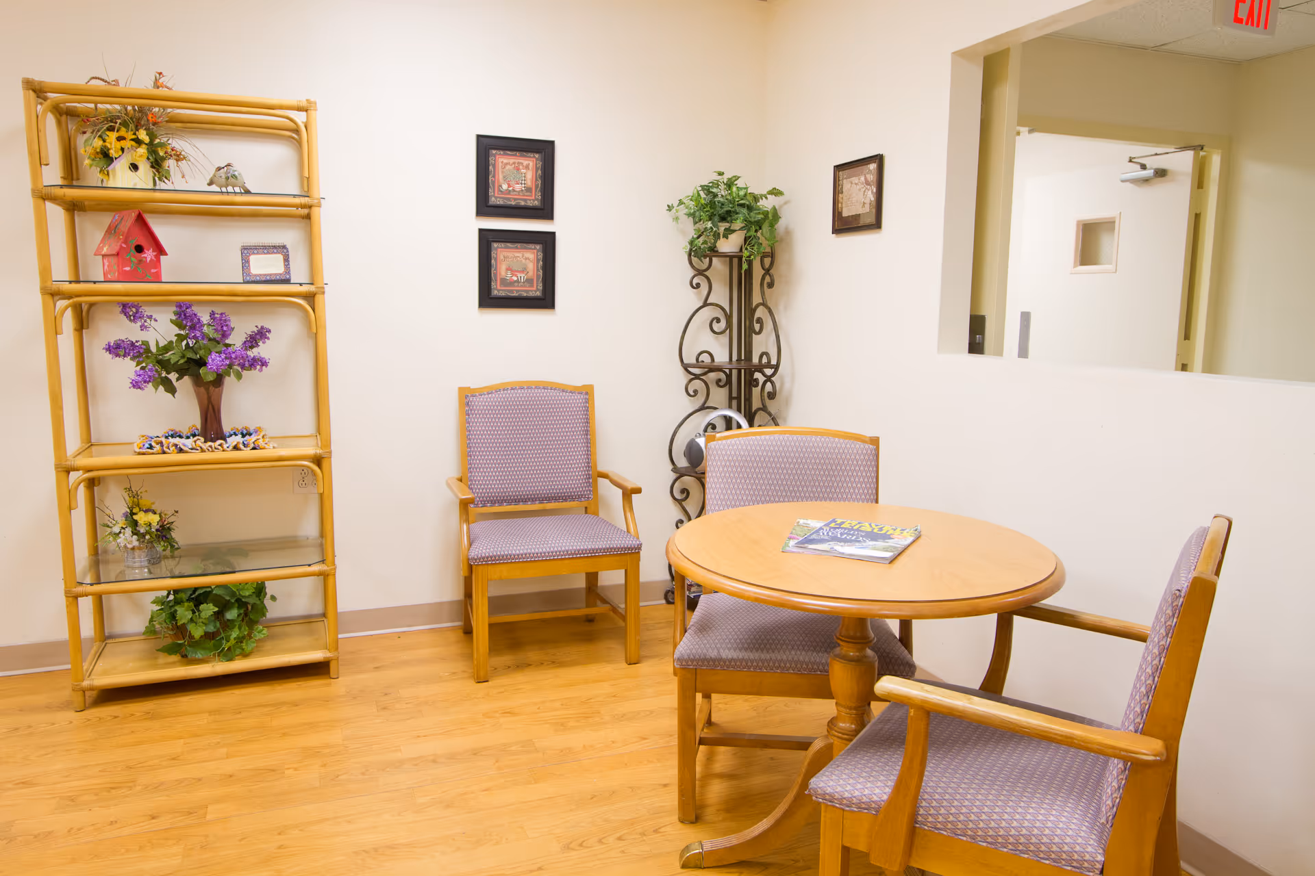 A small seating area with a round wooden table surrounded by three wooden chairs with purple patterned cushions. To the left, there is a tall bamboo shelf with decorative items including flowers and a small red birdhouse. On the wall behind the chairs, there are three framed pictures and a decorative metal plant stand with a green plant. The floor is wooden, and there is a window-like opening in the wall showing a hallway with a door and an exit sign above it.