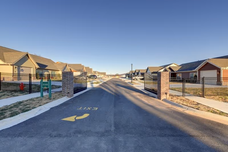 View of a gated entrance to a residential community with single-story houses on both sides of a paved road under a clear blue sky.