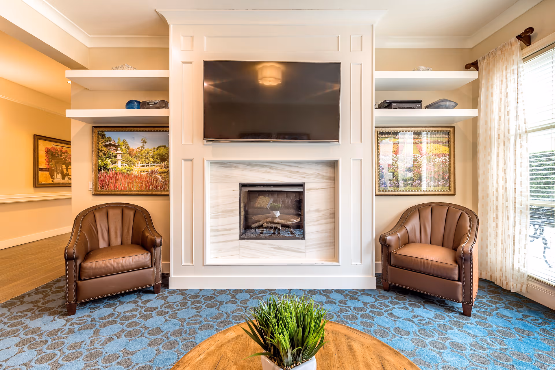 A cozy senior living room area featuring a white fireplace with a mounted flat-screen TV above it. Two brown leather armchairs are positioned on either side of the fireplace. The room has blue patterned carpet, a round wooden coffee table with a small green plant, and large windows with sheer curtains allowing natural light to enter. Framed landscape paintings are hung on the walls beside the fireplace.
